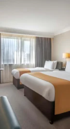 Bright hotel room with twin beds, cosy seating, and natural light through a large window.