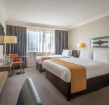 Bright hotel room with twin beds, cosy seating, and natural light through a large window.