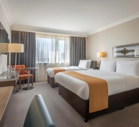 Bright hotel room with twin beds, cosy seating, and natural light through a large window.