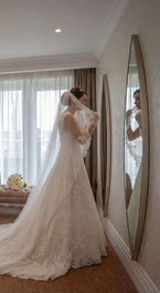 Bride in elegant suite adjusting veil next to a full-length mirror, soft lighting in a cosy room.