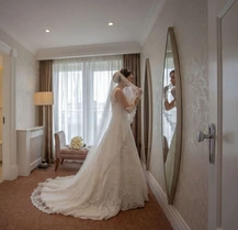 Bride in elegant suite adjusting veil next to a full-length mirror, soft lighting in a cosy room.
