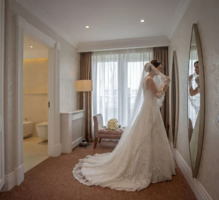 Bride in elegant suite adjusting veil next to a full-length mirror, soft lighting in a cosy room.