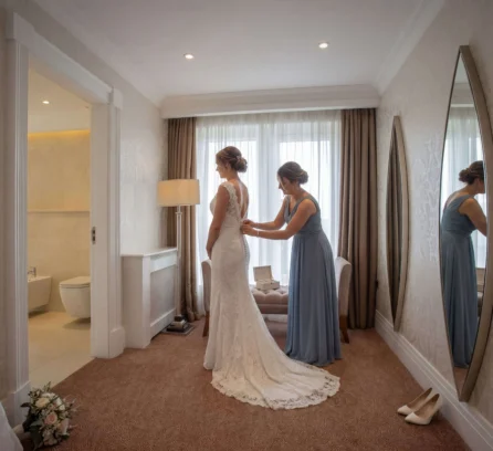 Bride in white gown with friend in hotel room, captured moments before a wedding.