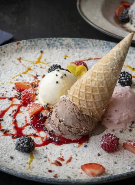 Three scoops of ice cream with fruit and cone on a speckled plate.