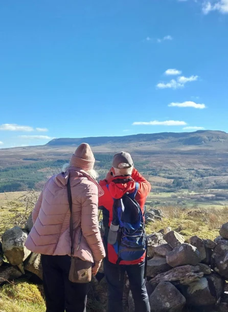 A couple in winter gear admire a scenic mountain view, framed by a rustic stone wall.