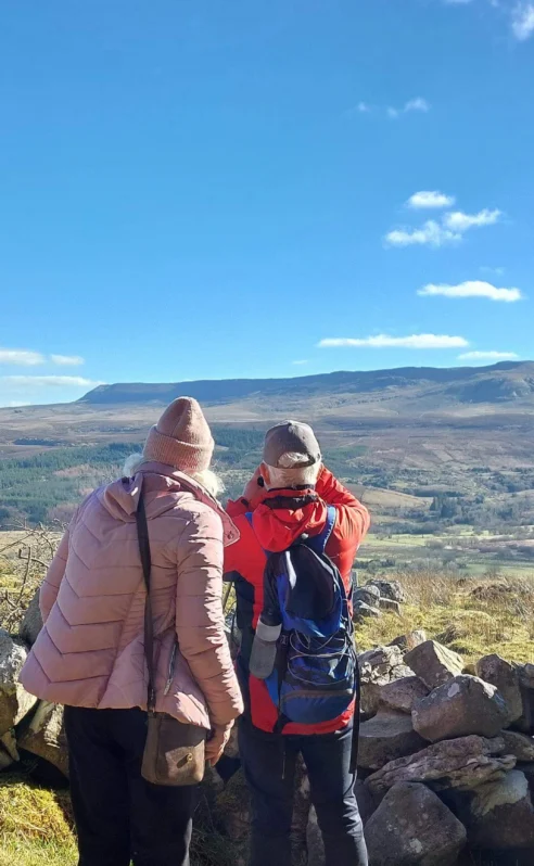 A couple in winter gear admire a scenic mountain view, framed by a rustic stone wall.