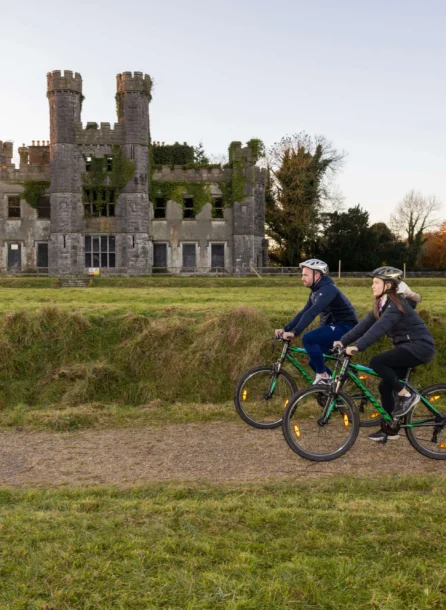 A couple cycling past a historic castle ruin in a lush, green landscape.