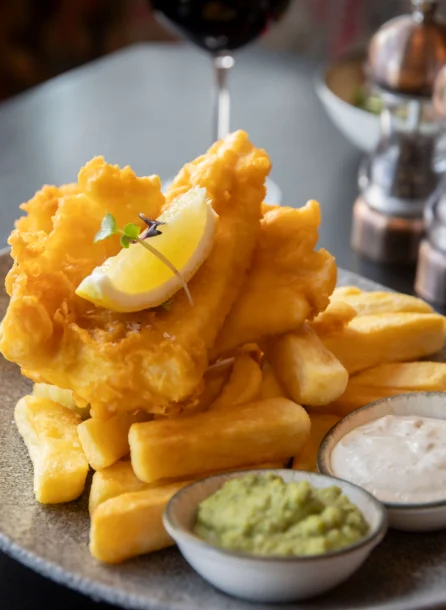 Plate of crispy fish and chips with tartar and mushy peas at a cosy restaurant setting.