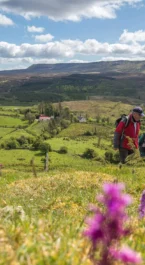 A couple hiking on a lush, green trail by Hotel Kilmore, with vibrant wildflowers and rolling hills under a blue sky.