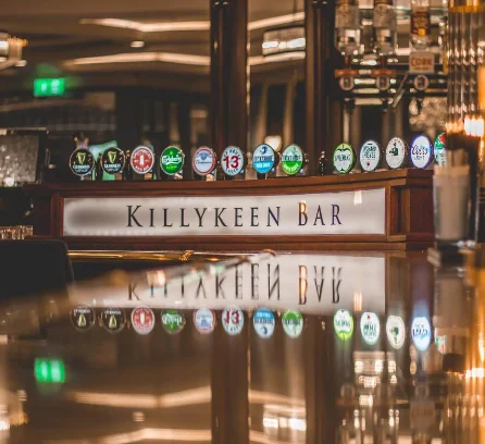 Warm, inviting bar with a polished counter and taps, labelled "Killykeen Bar". Quiet ambiance.