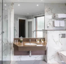 Modern hotel bathroom with marble tiles, glass shower, and neatly arranged toiletries.