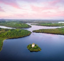 Serene landscape with lush greenery, calm waters, and a historic tower under a pink sunset sky.