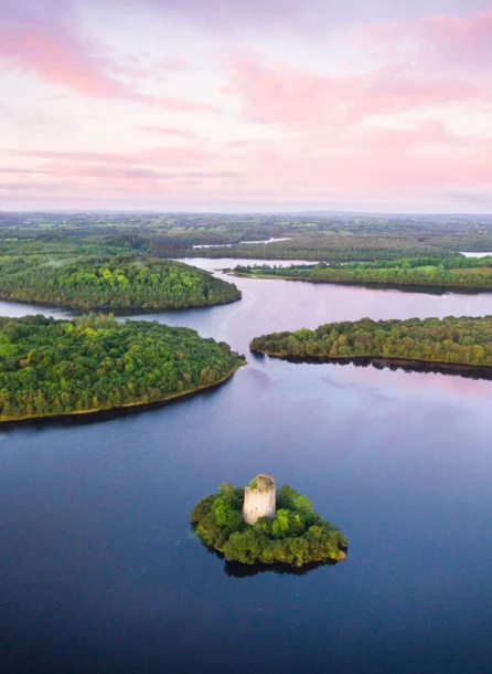 Serene landscape with lush greenery, calm waters, and a historic tower under a pink sunset sky.