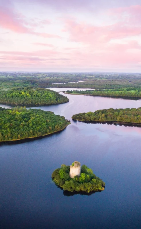 Serene landscape with lush greenery, calm waters, and a historic tower under a pink sunset sky.